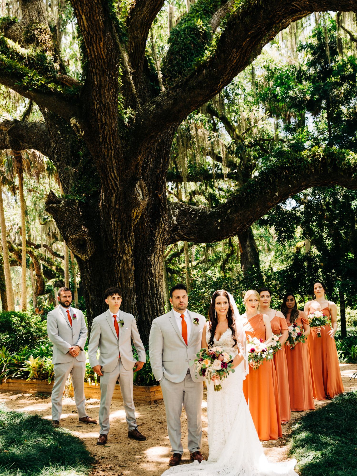Wedding party poses under a majestic oak tree with bridesmaids in coral dresses and groomsmen in gray suits.
