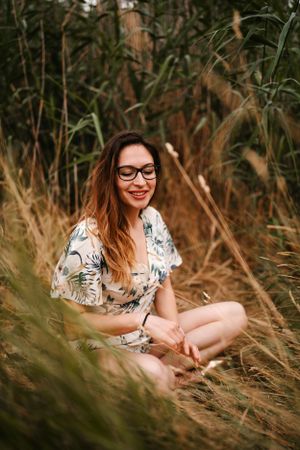 A woman in a floral dress sits in tall grass during a summer photoshoot.