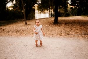 A small child in a white dress stands alone on a dirt path in a wooded area.