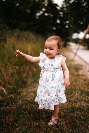 A toddler in a floral dress smiles while walking outdoors in tall grass.