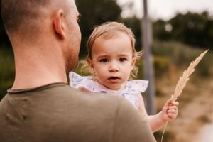 A close-up portrait of a toddler in a pink dress holding wheat grain while being held on a rural path.