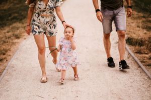 A toddler in a floral dress takes early steps on a path while holding hands with parents.