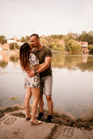 Couple embracing by a tranquil lake during sunset with lakeside homes in the background.