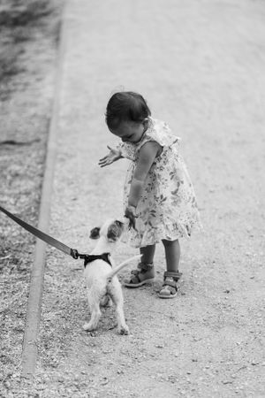 A black and white photo of a small child in a dress petting a small dog on a path.