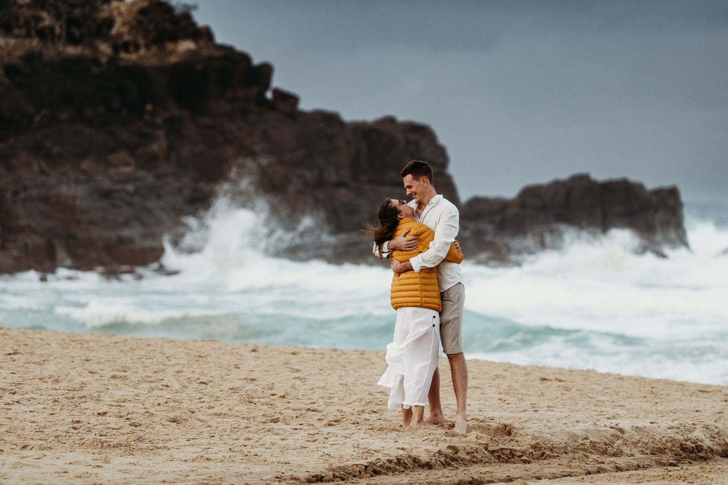 A couple embraces on a beach with dramatic rocky cliffs and crashing waves in the background.