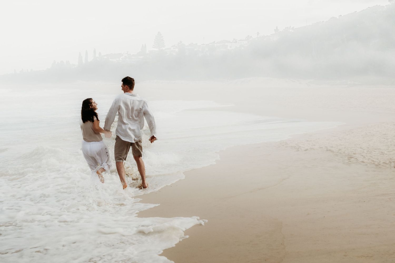 Two people walking together through shallow ocean waves on a foggy beach.