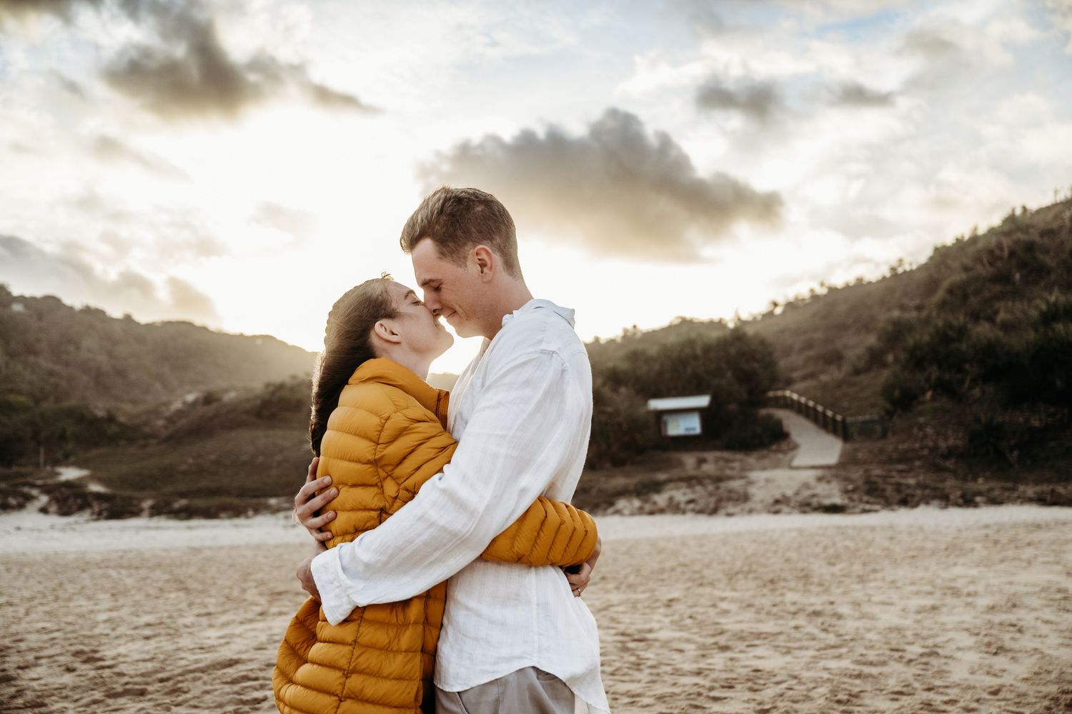 A romantic couple in white and yellow clothing embrace against a sunset mountain backdrop.
