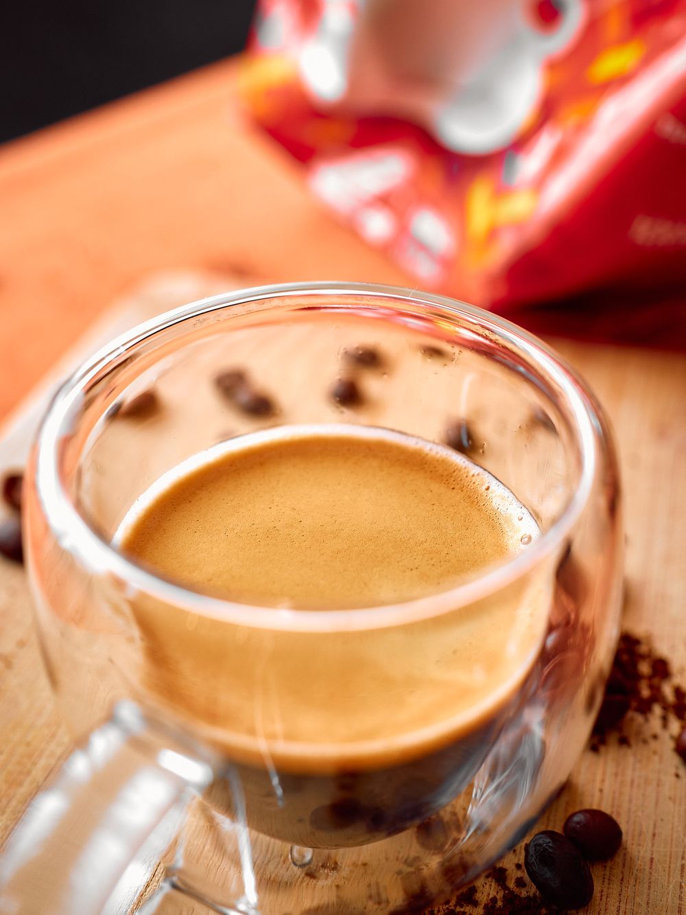 Close-up of a fresh espresso shot in a clear glass cup with coffee beans scattered around.