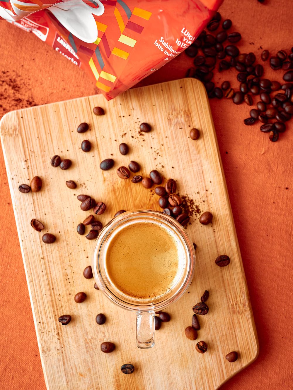 Overhead view of espresso in a glass cup surrounded by coffee beans on a wooden cutting board.