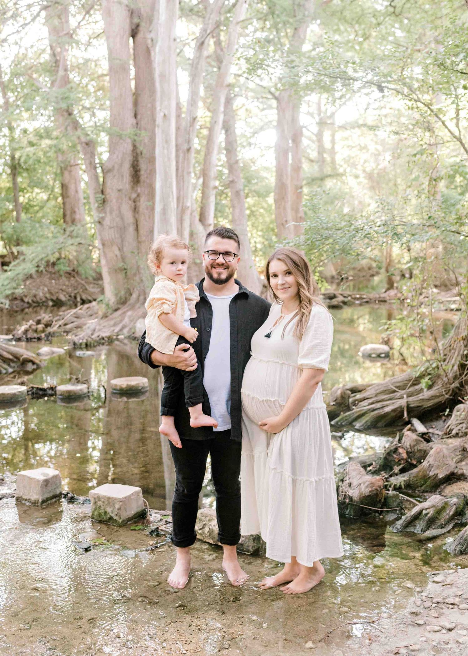 A family stands together by a creek in the woods wearing coordinated outfits for maternity photos.