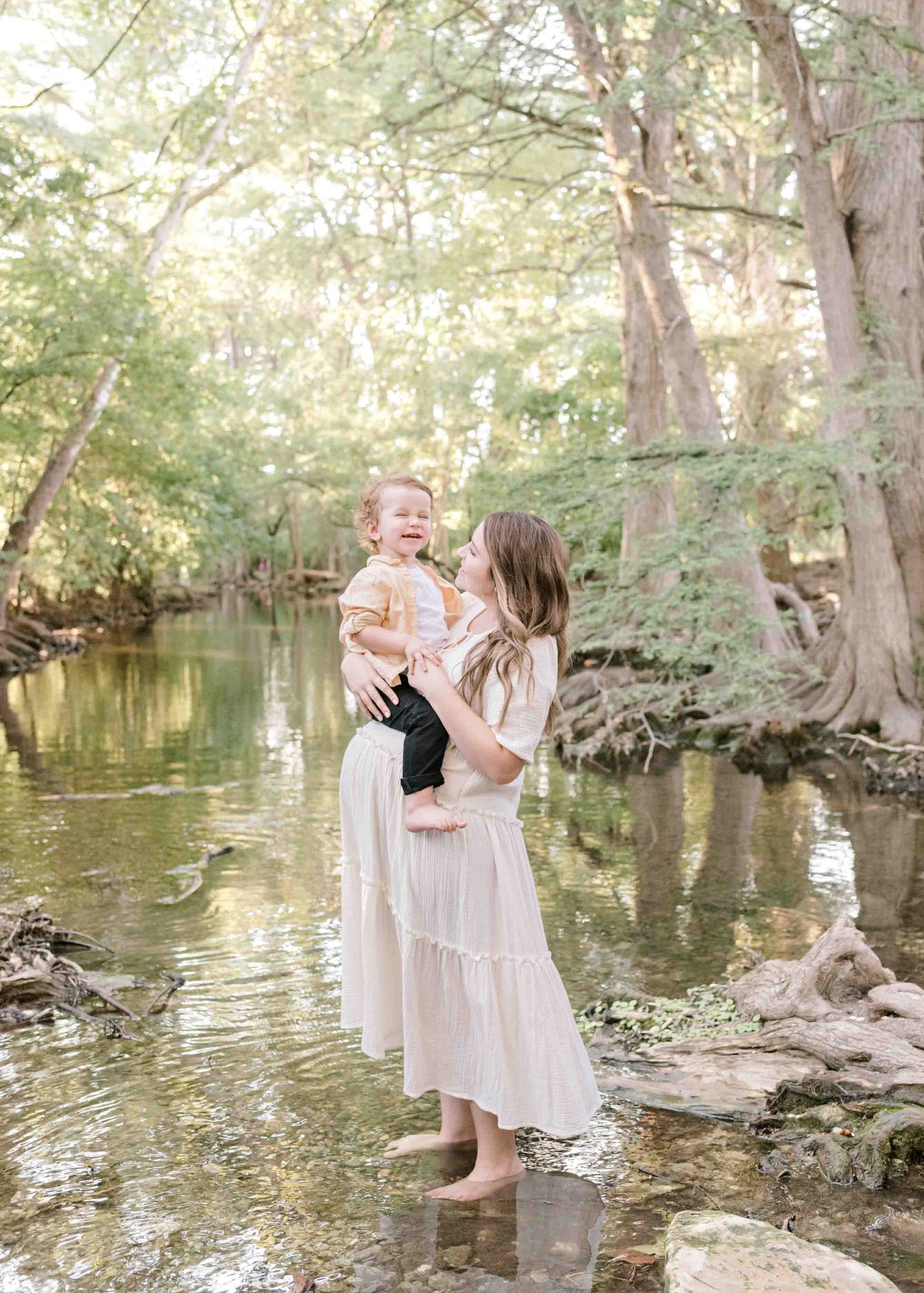 Mother and child share a joyful moment standing in a sunlit creek surrounded by trees.
