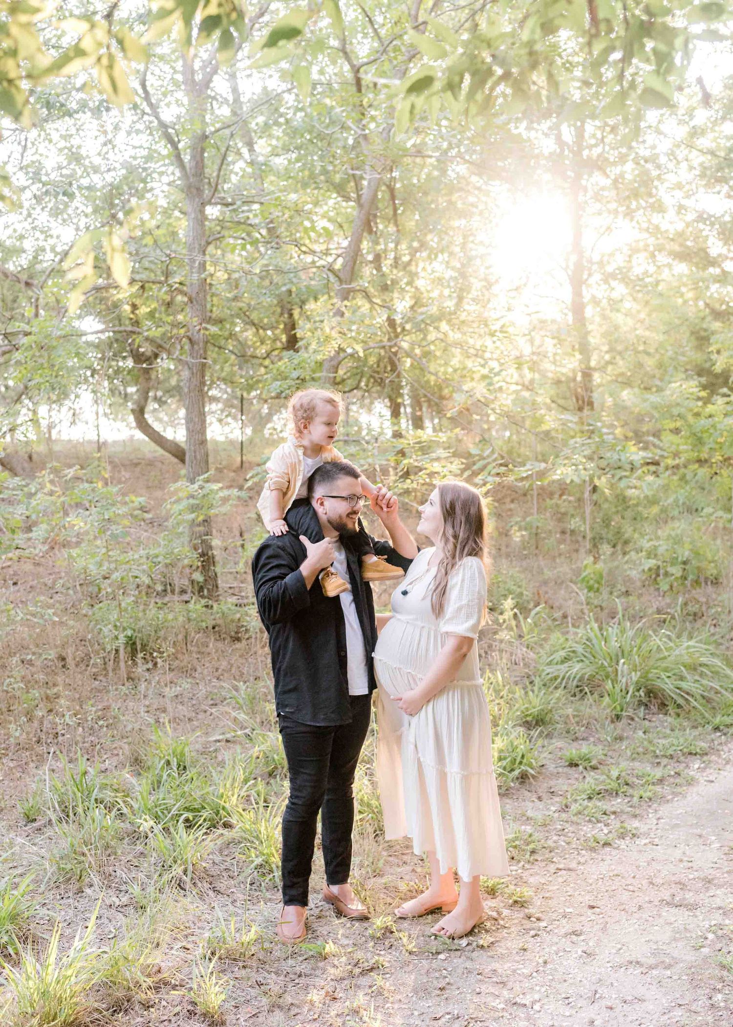 Family maternity photoshoot in a sun-dappled forest setting with golden light streaming through trees.