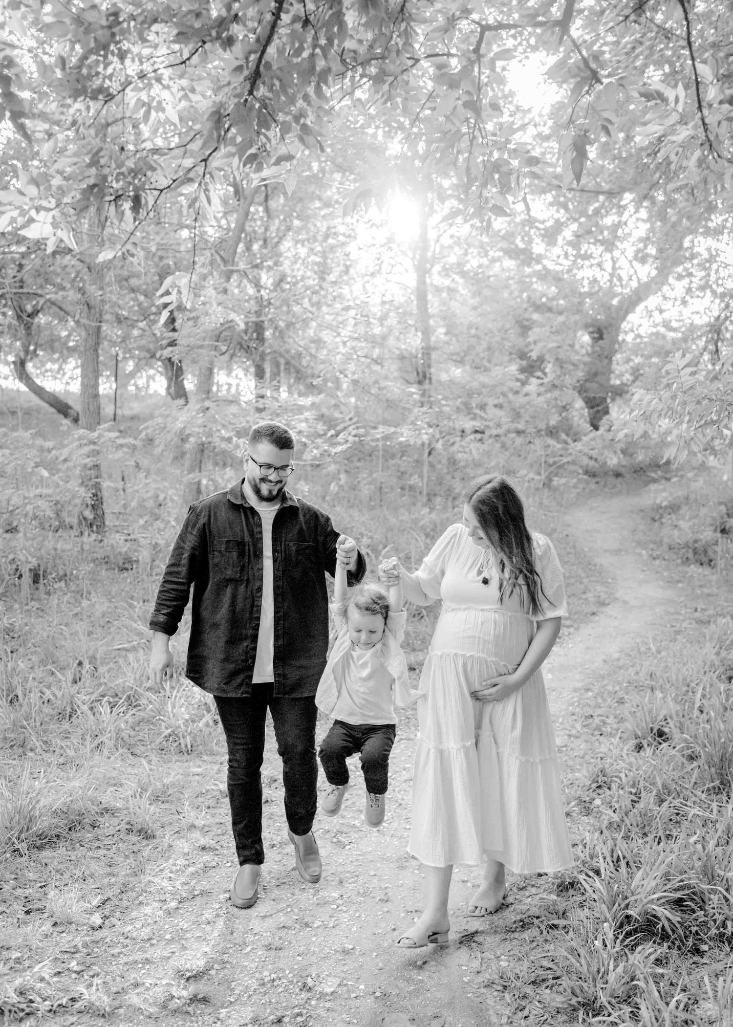 A family enjoys a sun-dappled walk together in a wooded area, with flowing white dress and casual attire.