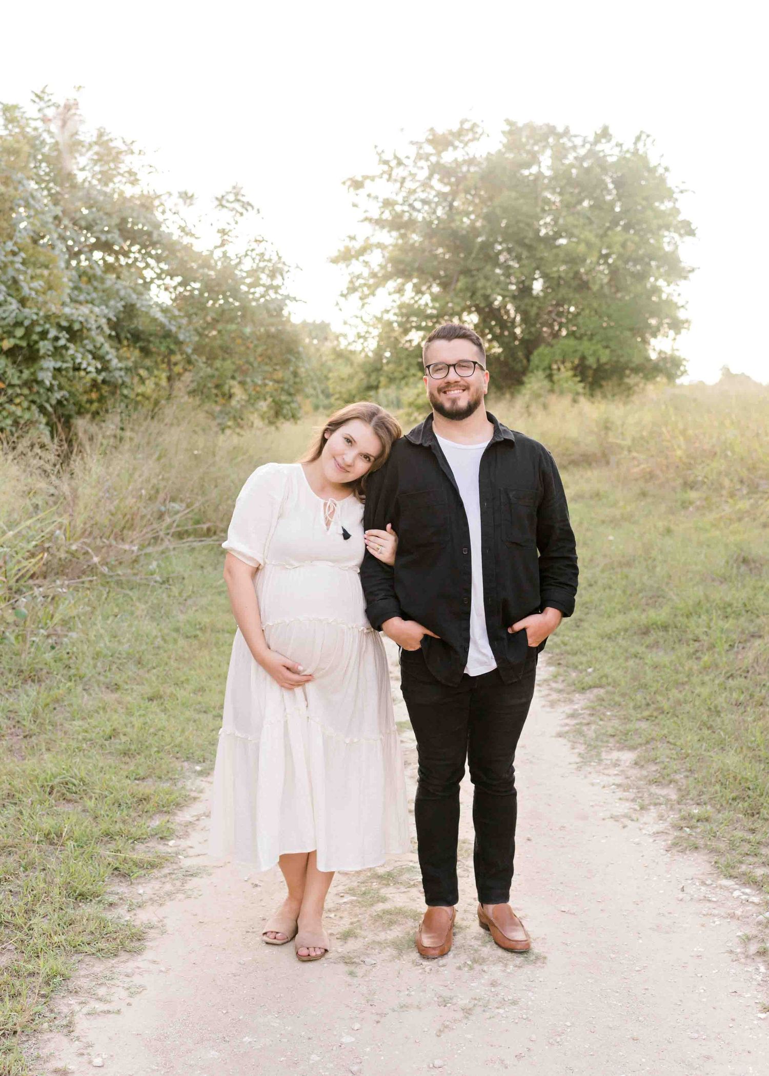 A couple poses on a dirt path during a sunset maternity photoshoot among blooming trees.