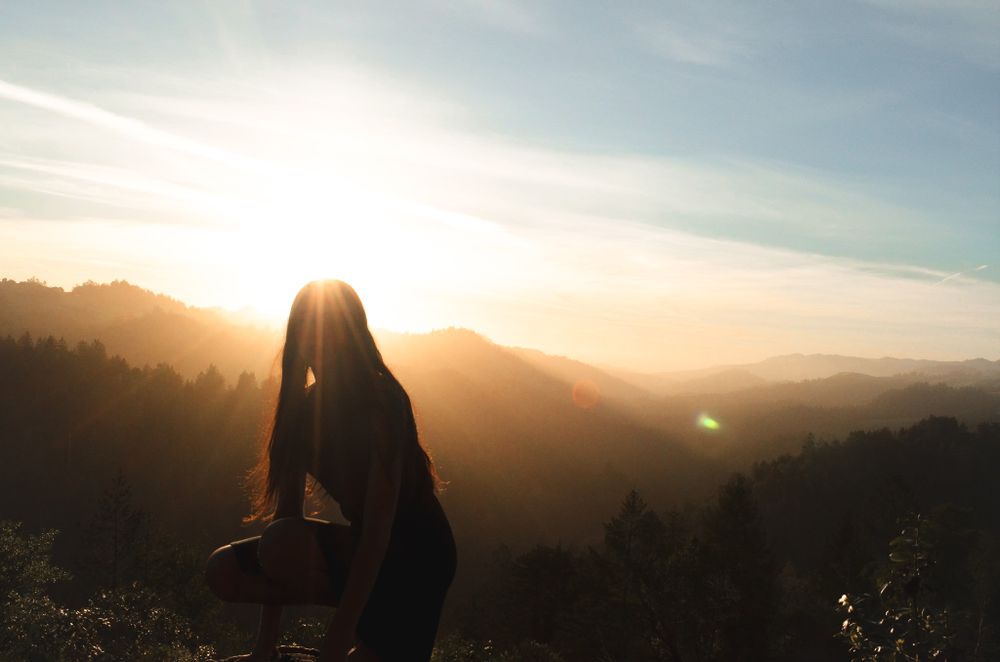 Silhouette standing on mountain peak during golden sunset with mountains in background.