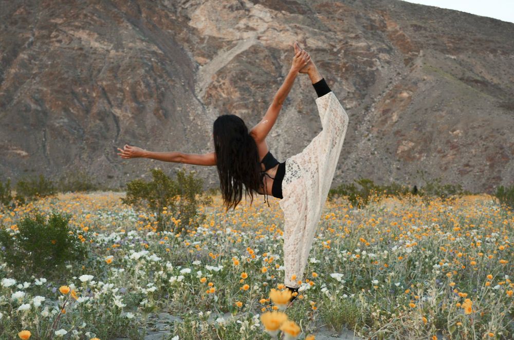 Person stretching gracefully in white outfit among wildflowers with mountains in background.