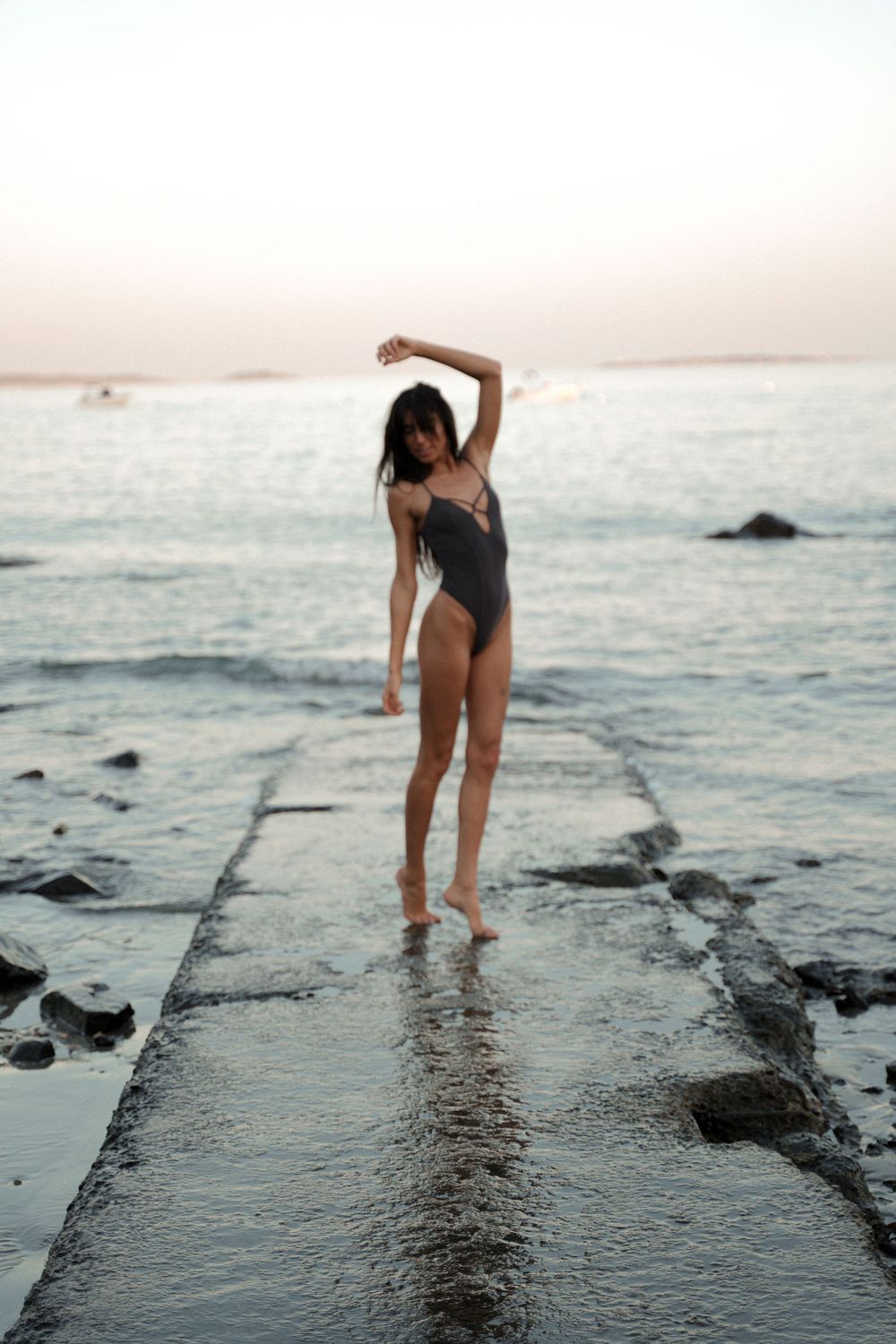 Silhouetted figure in swimsuit stands on wet jetty against ocean backdrop.