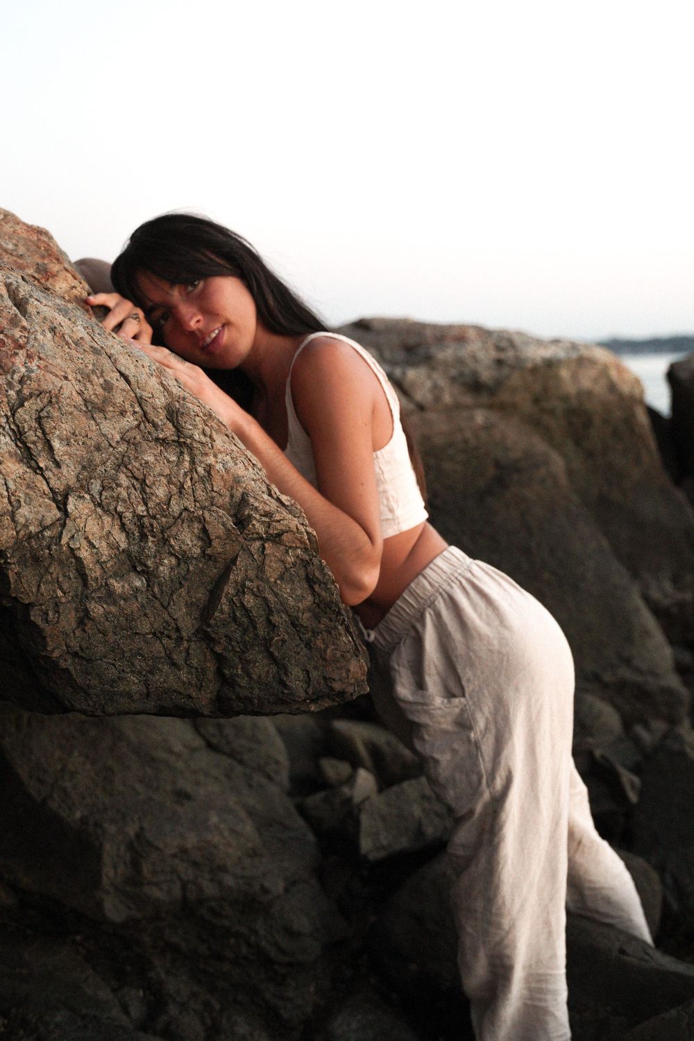 Figure leaning against rugged coastal rocks in white outfit at dusk.