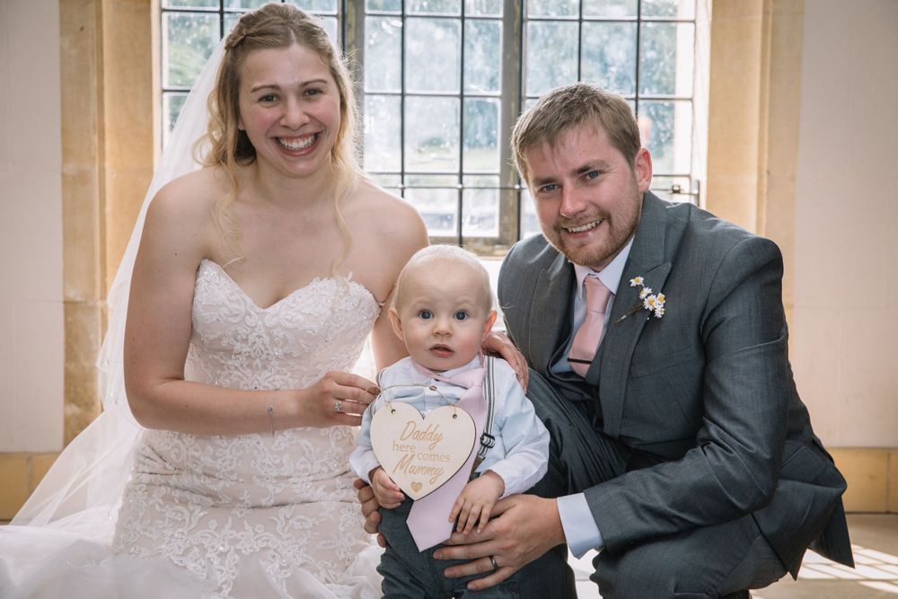 A happy wedding couple poses with their infant child by a bright window.