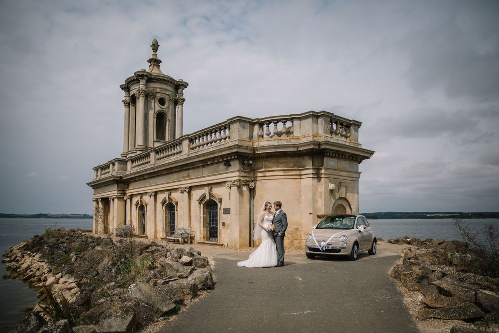 Newlyweds pose outside a historic church overlooking the water with their wedding car.