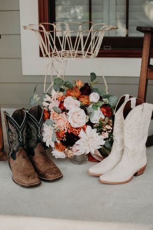 Rustic wedding setup featuring cowboy boots, floral arrangement and white metal basket on porch.