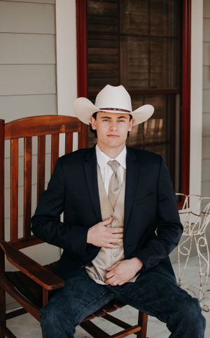 A person in western formal wear sitting on a wooden rocking chair on a porch.