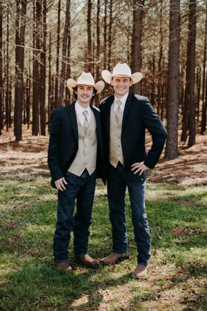 Two people in matching formal western attire and white cowboy hats pose in a forest setting.