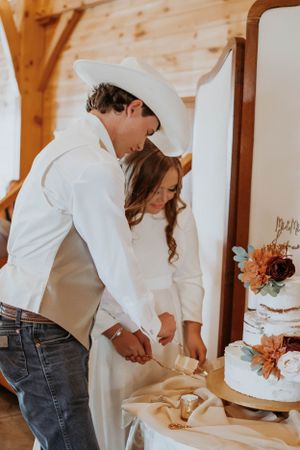 Newlyweds cut their elegant three-tier white wedding cake together at a rustic venue.