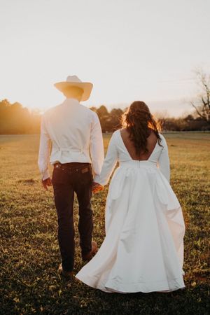 Bride in backless dress and groom in cowboy hat walking together at sunset in an open field.