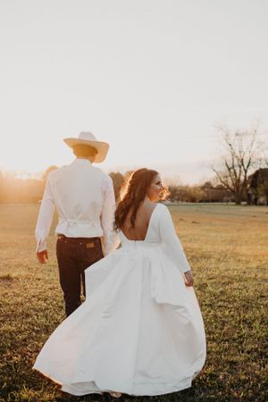 Newlywed couple walking in a field at golden hour, bride in flowing white dress and groom in white shirt and cowboy hat.