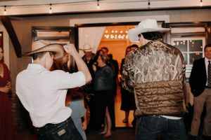 Two people in cowboy hats dance energetically at an indoor party with string lights and onlookers.