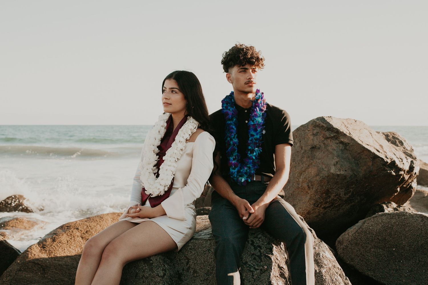 A brother and sister wear graduation leis while sitting at rocks at the beach.