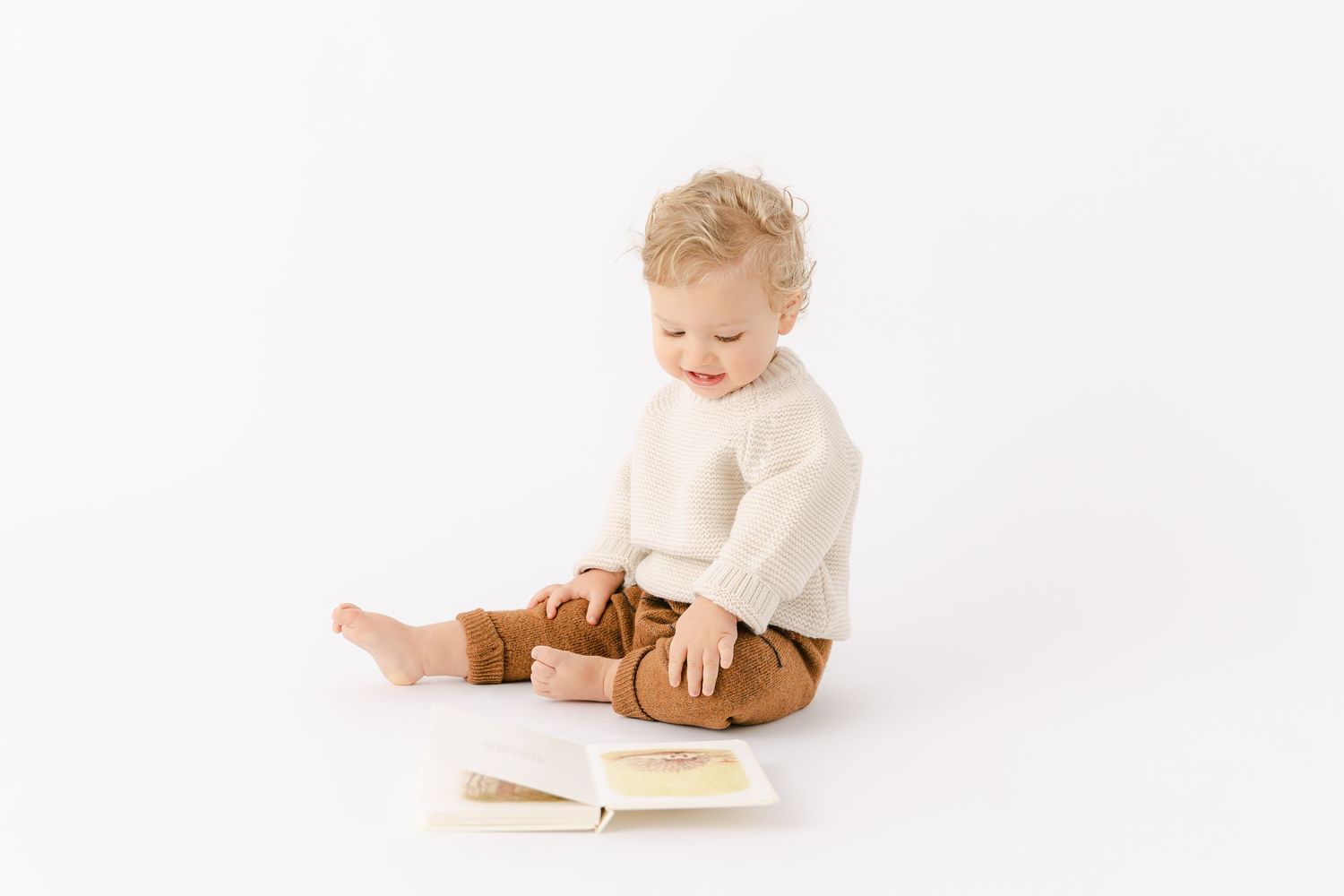 One year old baby with curly blonde hair wears a cream knit sweater and brown knit pants and looks down at a book.
