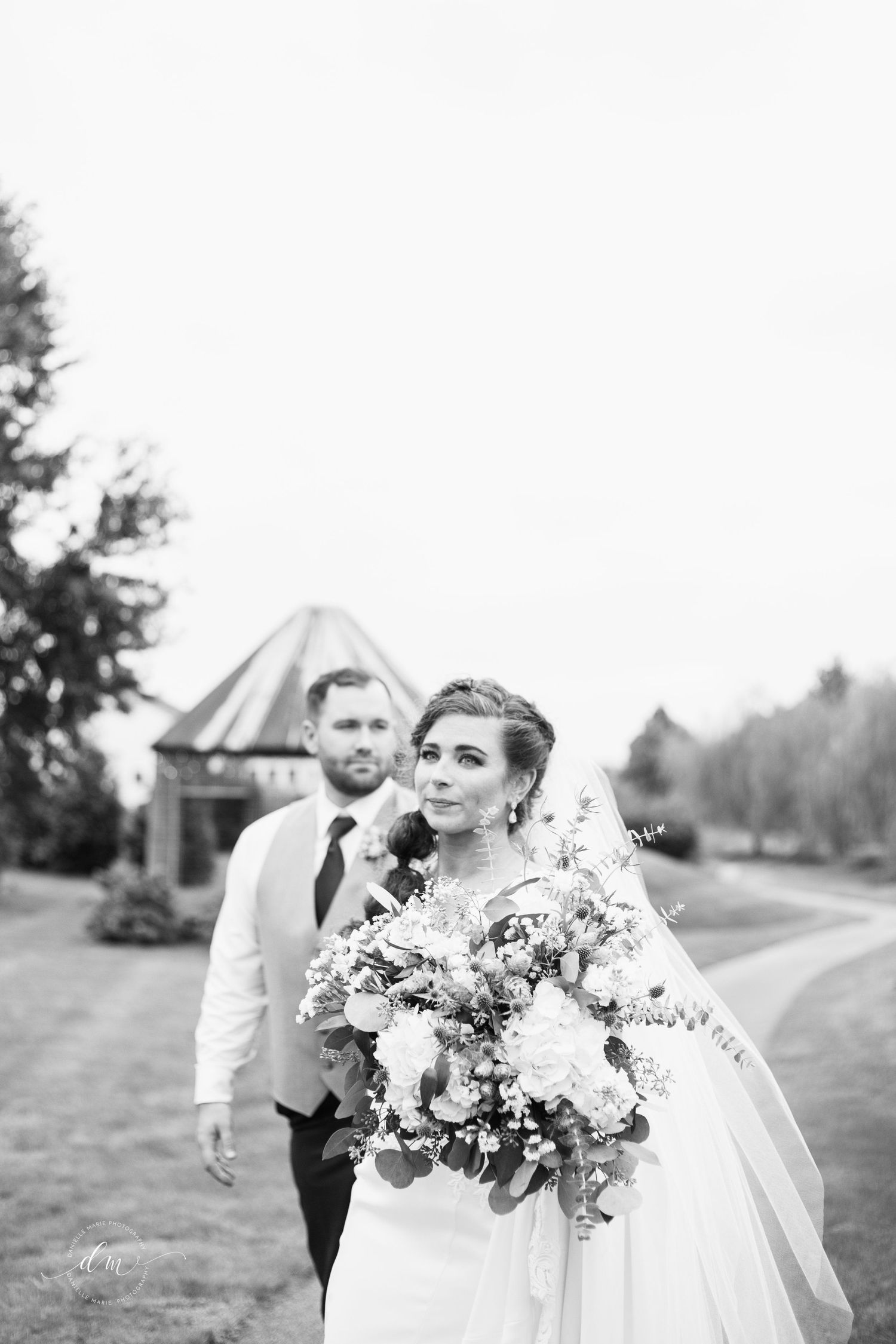 Black and white wedding portrait of a bride holding a bouquet with groom in background.