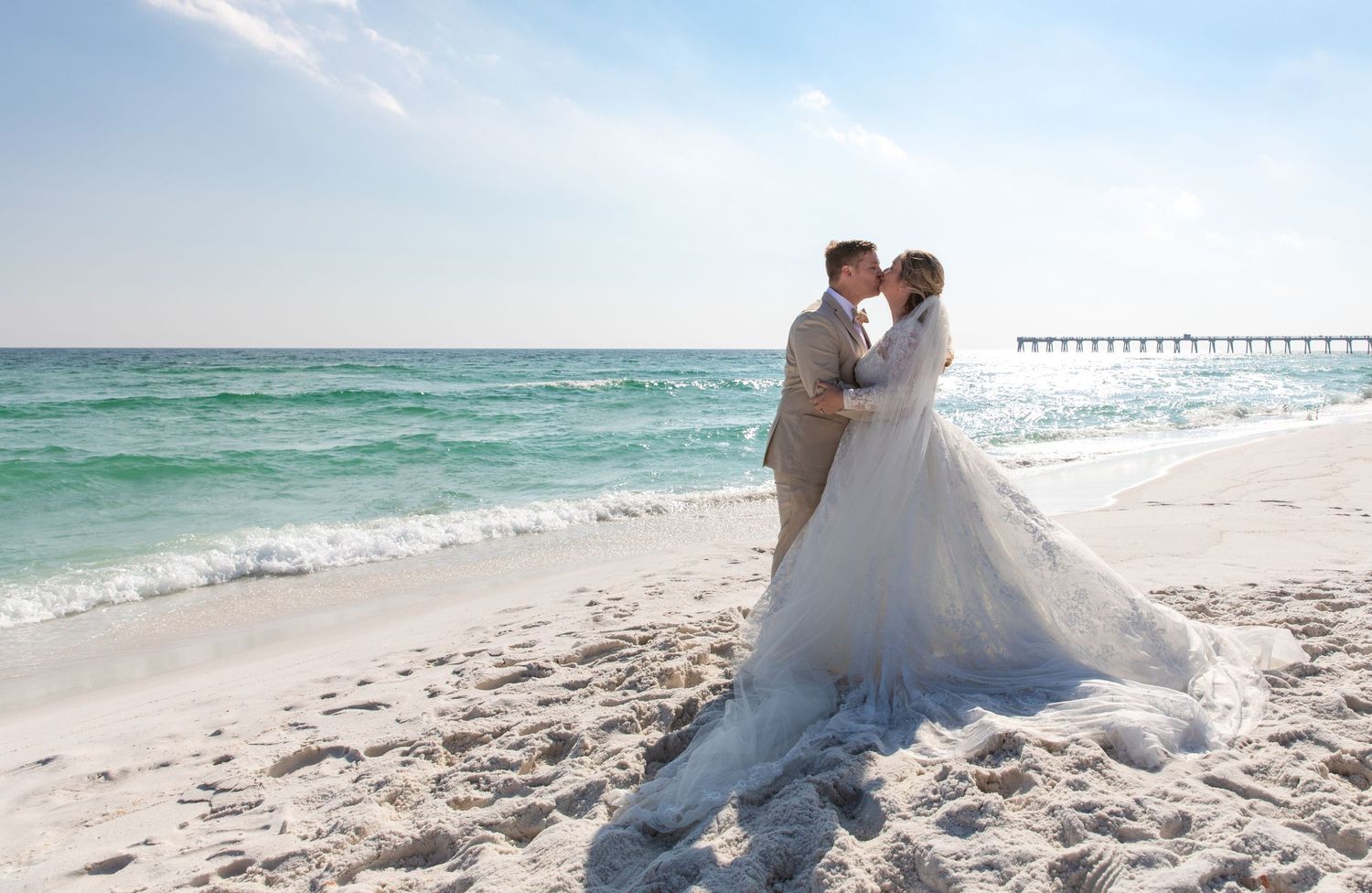 A couple in wedding attire embraces on a white sandy beach with a pier visible in the background.