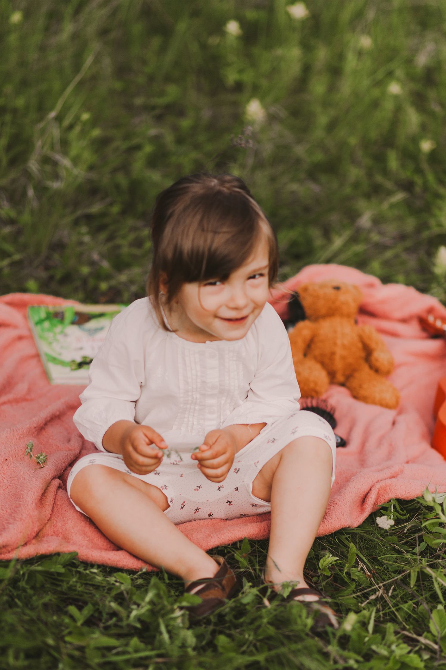 Child in white dress sitting on coral blanket playing in grass outdoors.