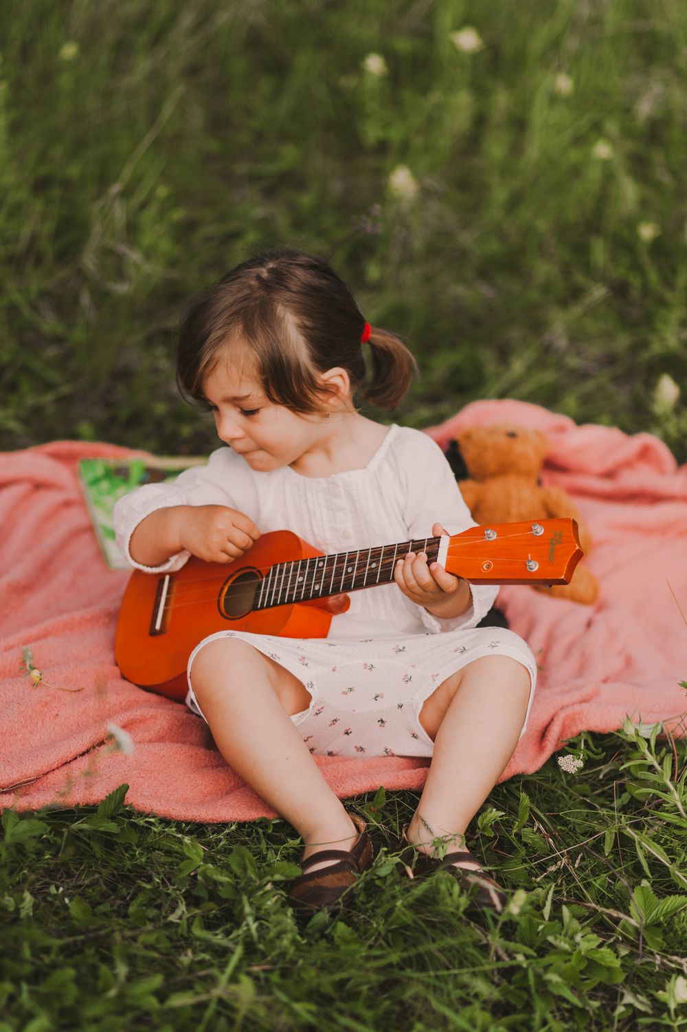 Child playing with orange ukulele while sitting on pink blanket in grassy field.
