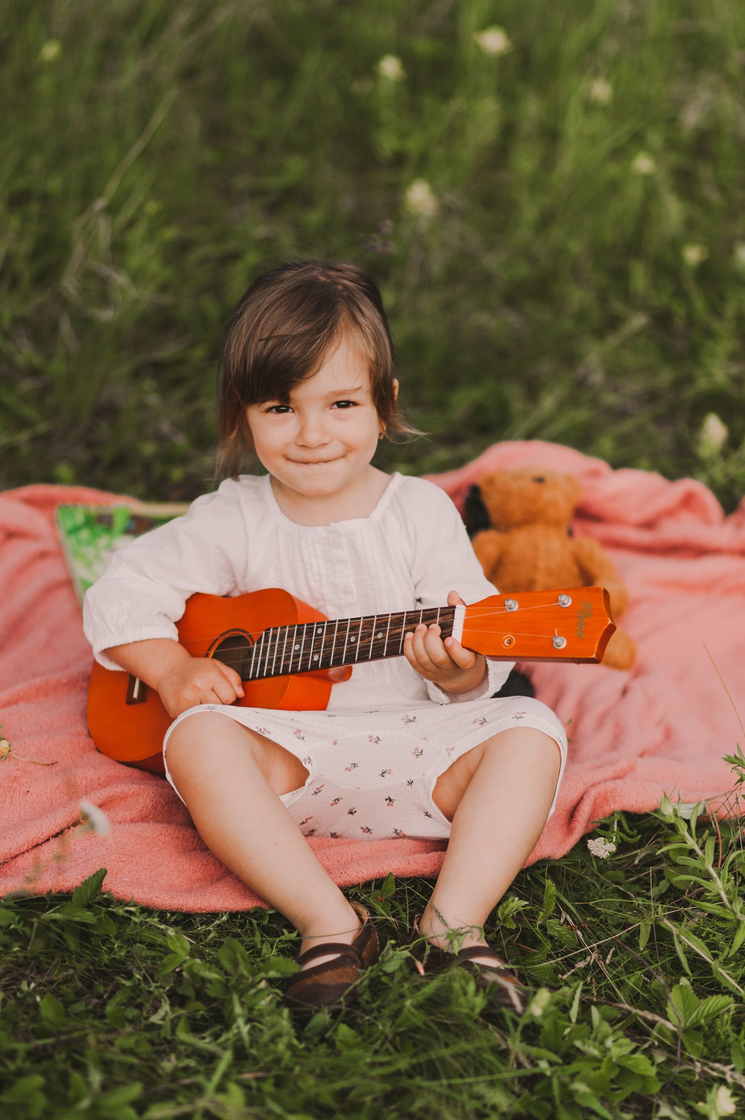 Young musician practicing ukulele while sitting on pink blanket outdoors.