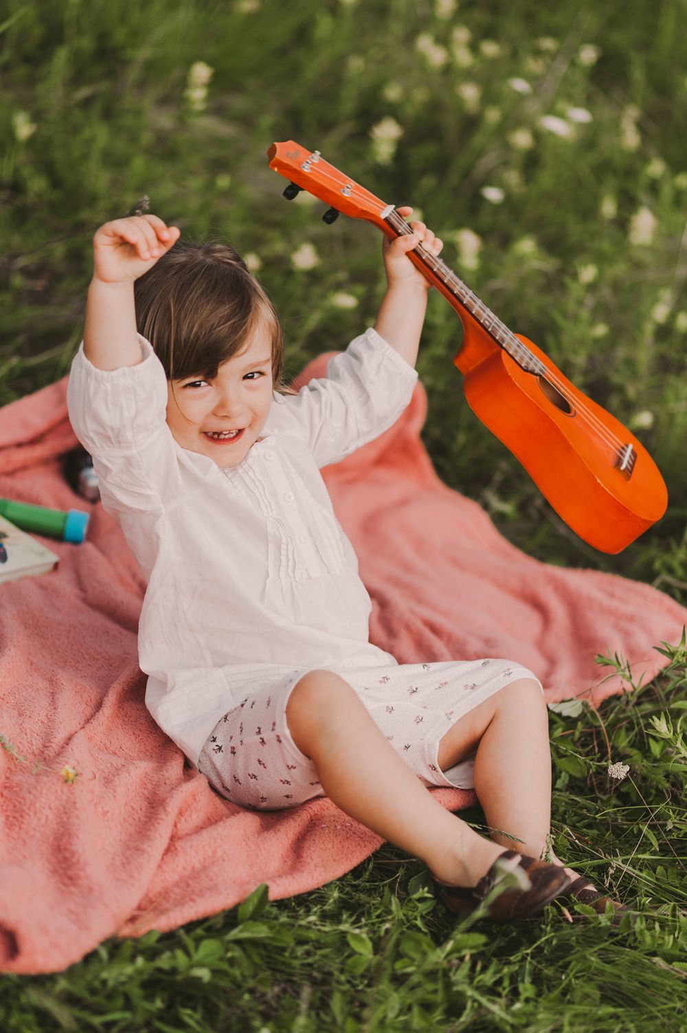Child in white dress joyfully playing with orange ukulele on pink blanket.