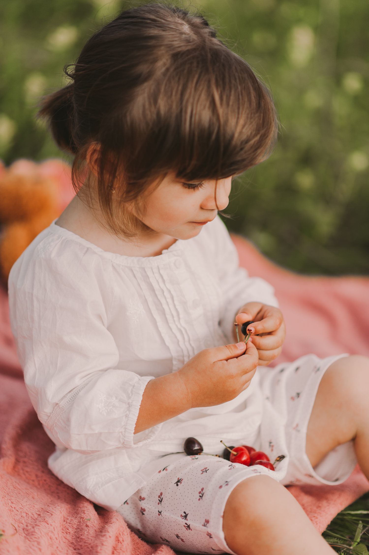 Child in white dress playing with toys on pink blanket in grass.