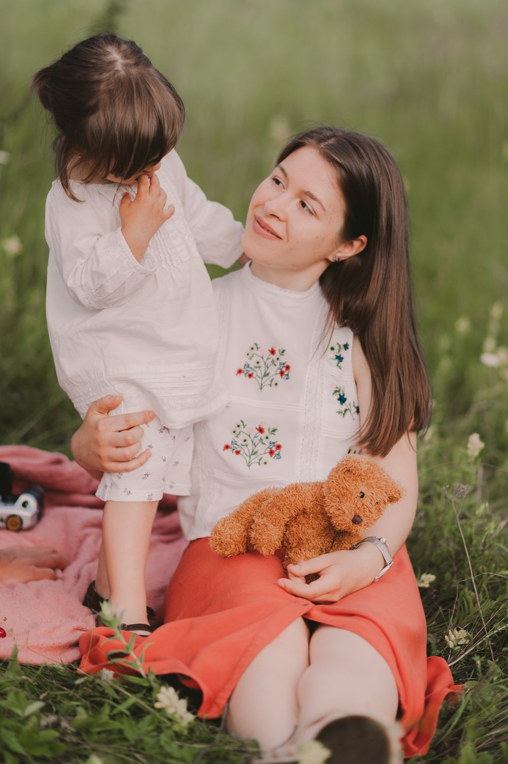 Parent and child sharing playful moment on coral blanket with teddy bear outdoors.