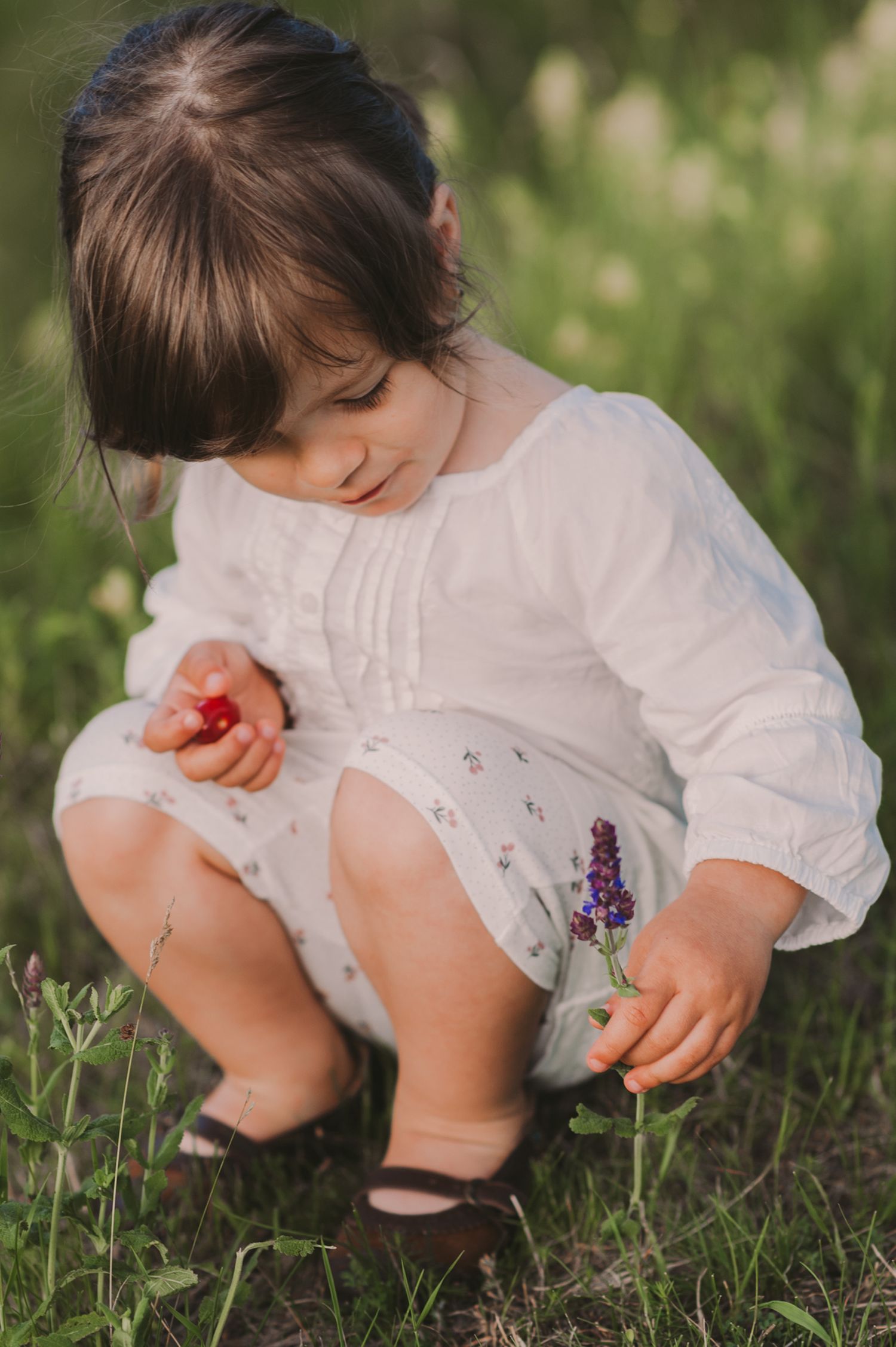 Child in white dress crouching down to explore flowers in a garden.