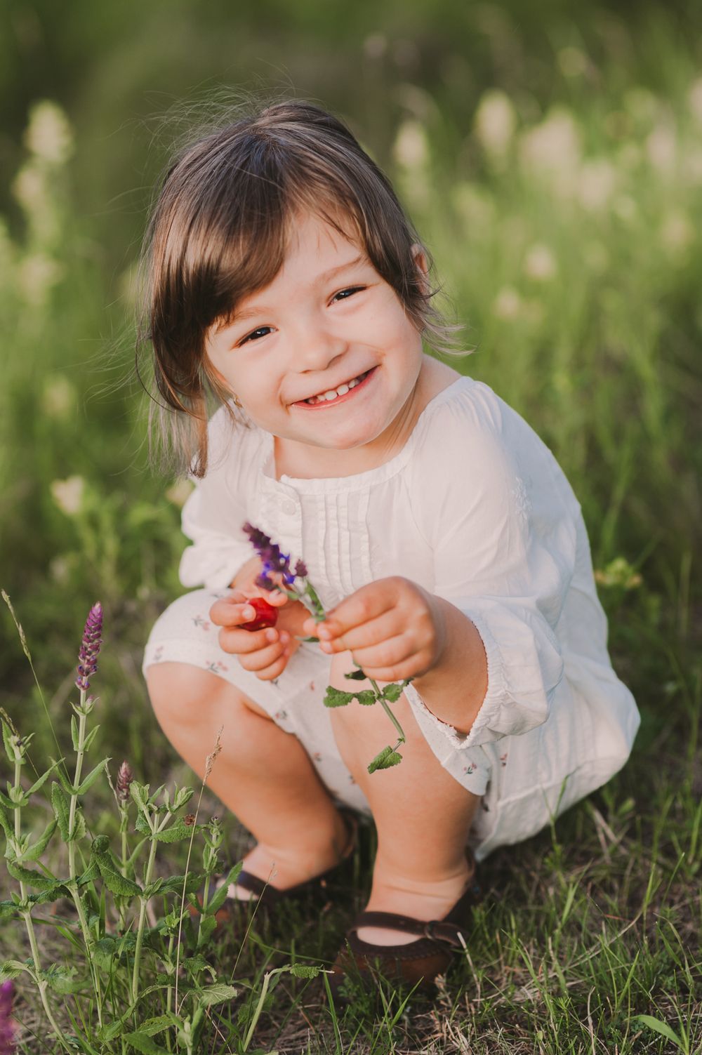 Child in white dress playing with flowers while crouching in green grass.