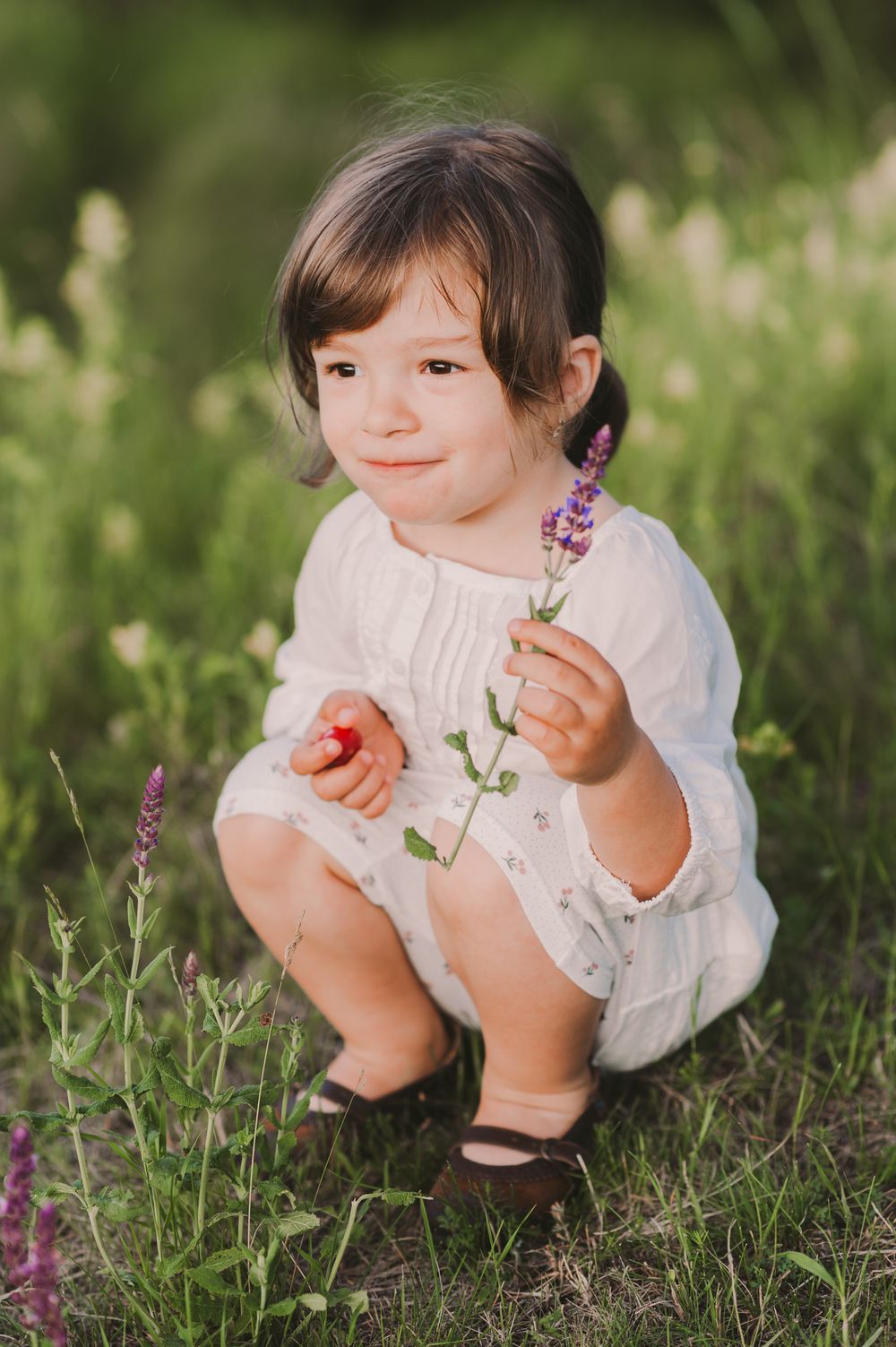 Child in white dress examining purple wildflowers in green grass.