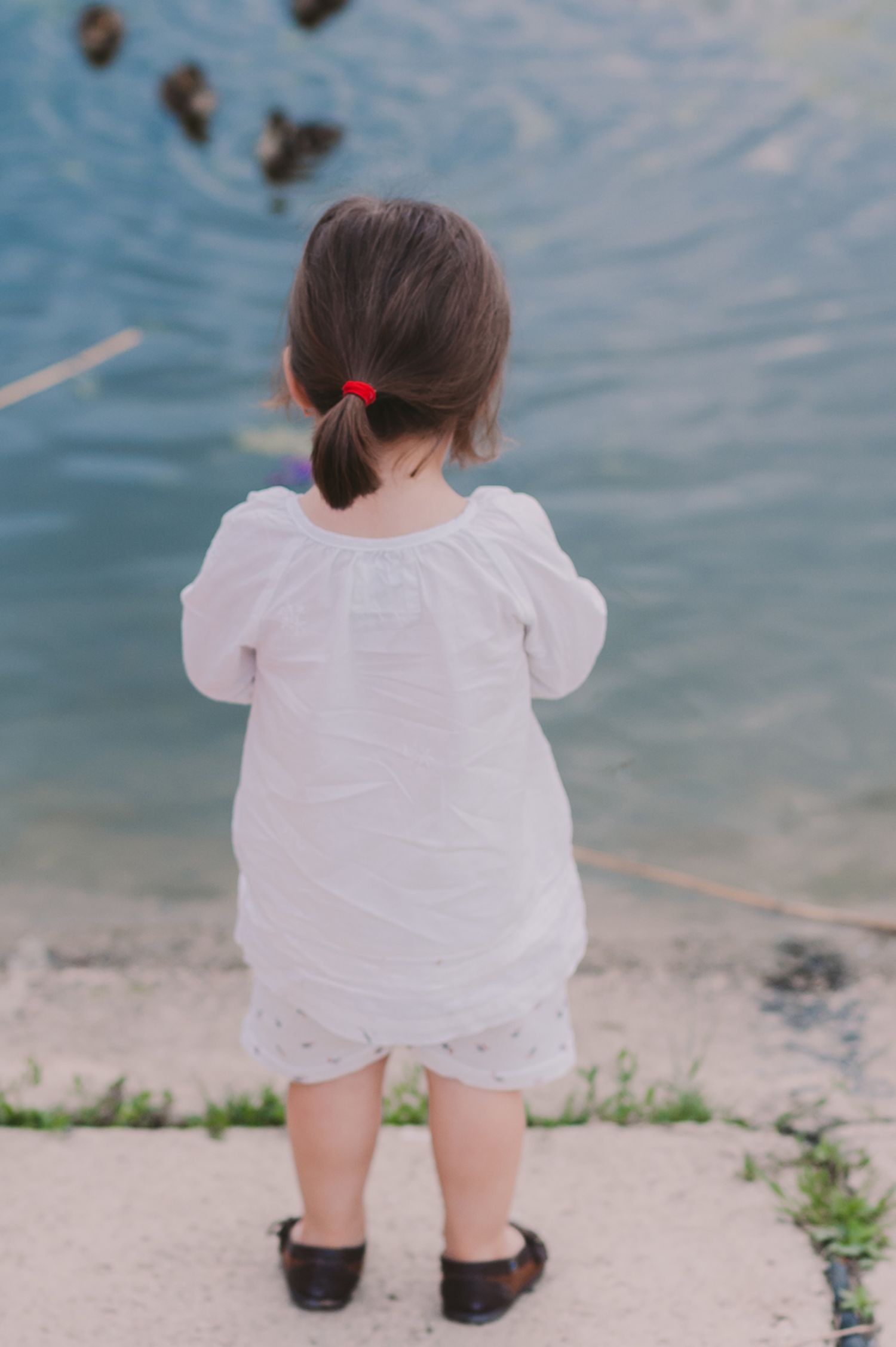 Child in white dress sitting by water's edge on a sunny day.
