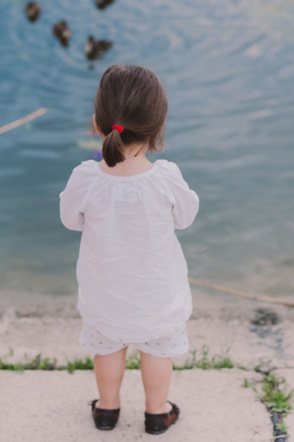 Child in white dress sitting by water's edge on a sunny day.