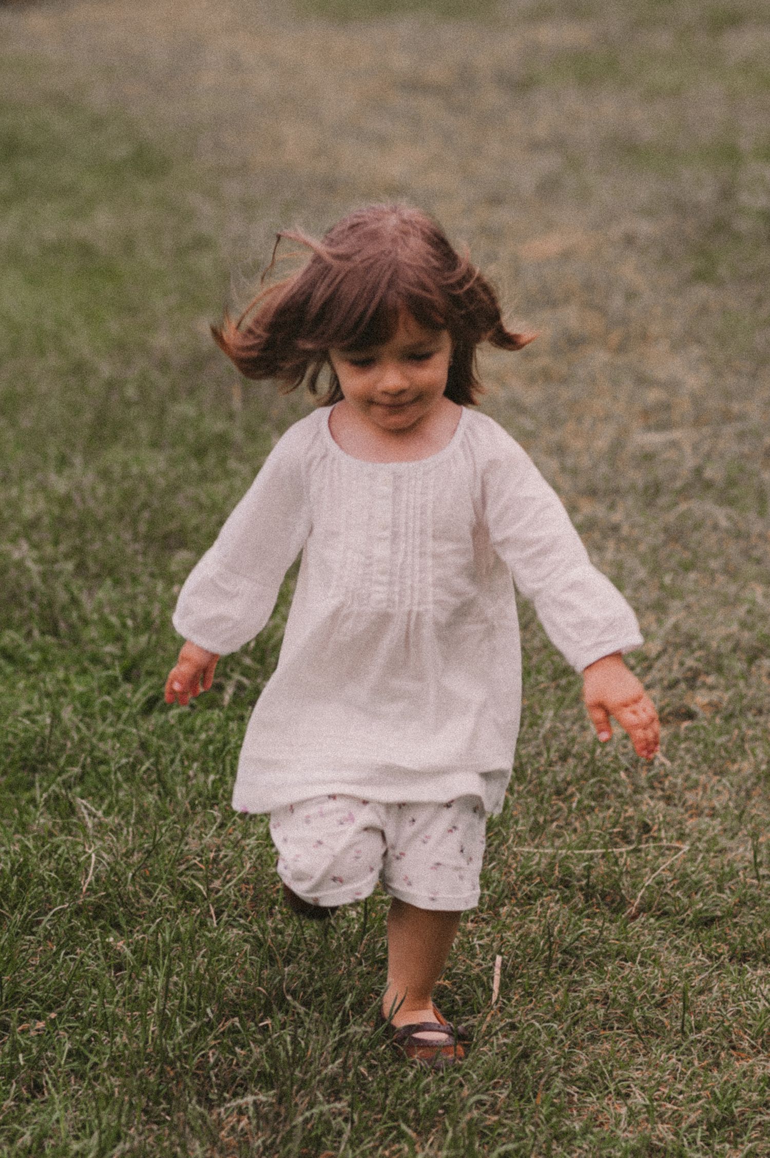 A toddler in a white dress explores a grassy meadow with joyful expression.