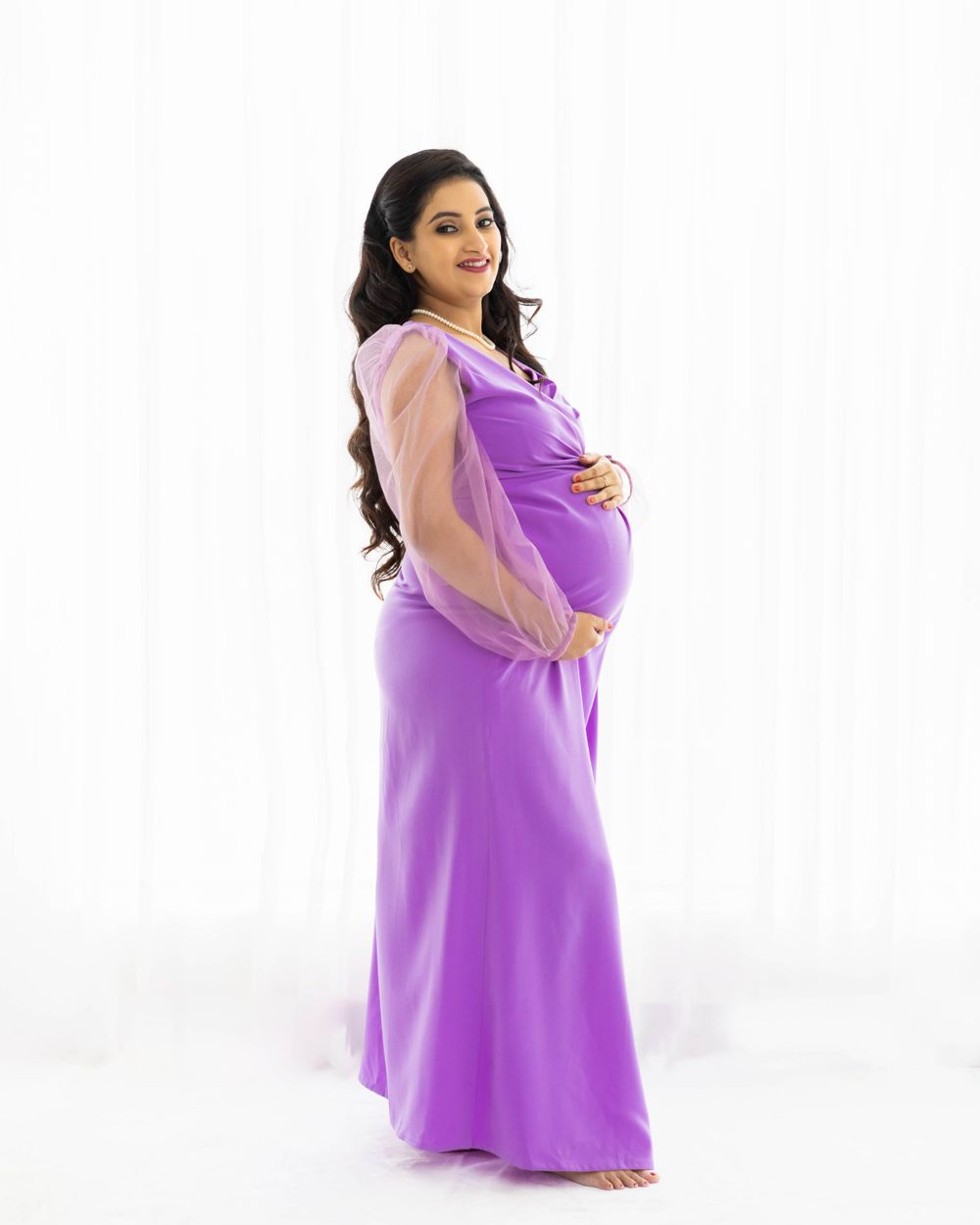 Maternity portrait featuring purple flowing dress with sheer sleeves against white backdrop.