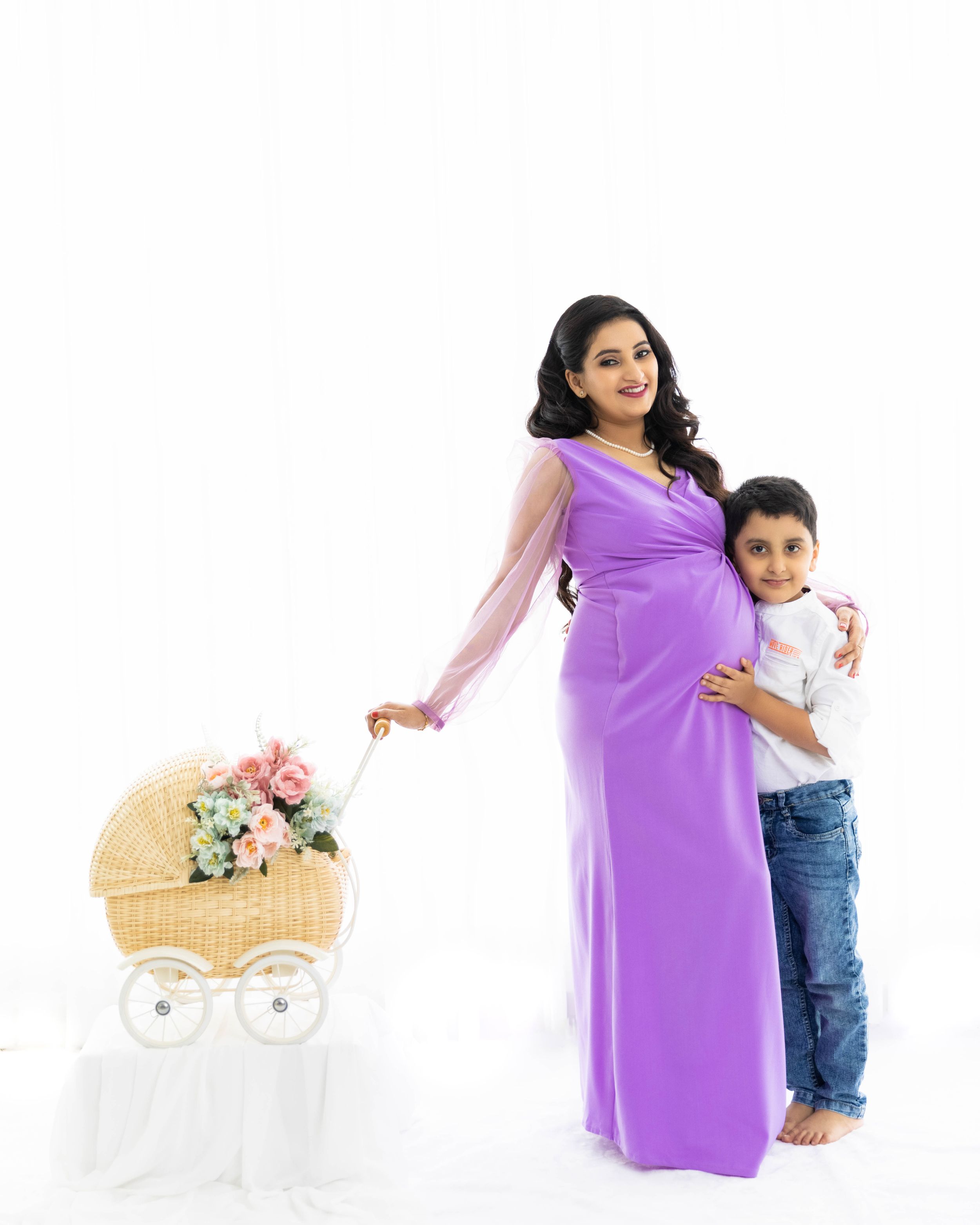 Maternity studio portrait with child and decorative baby carriage against white background.
