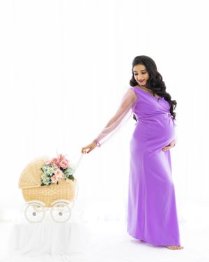 Pregnant woman in purple dress poses with decorative baby carriage prop.